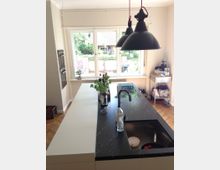 Moderne Küche mit großer Kücheninsel, die eine schwarze Arbeitsplatte und ein eingebautes Spülbecken besitzt. Im Hintergrund befindet sich ein großes Fenster mit Blick auf einen Garten, und zwei hängende schwarze Lampen beleuchten den Raum.