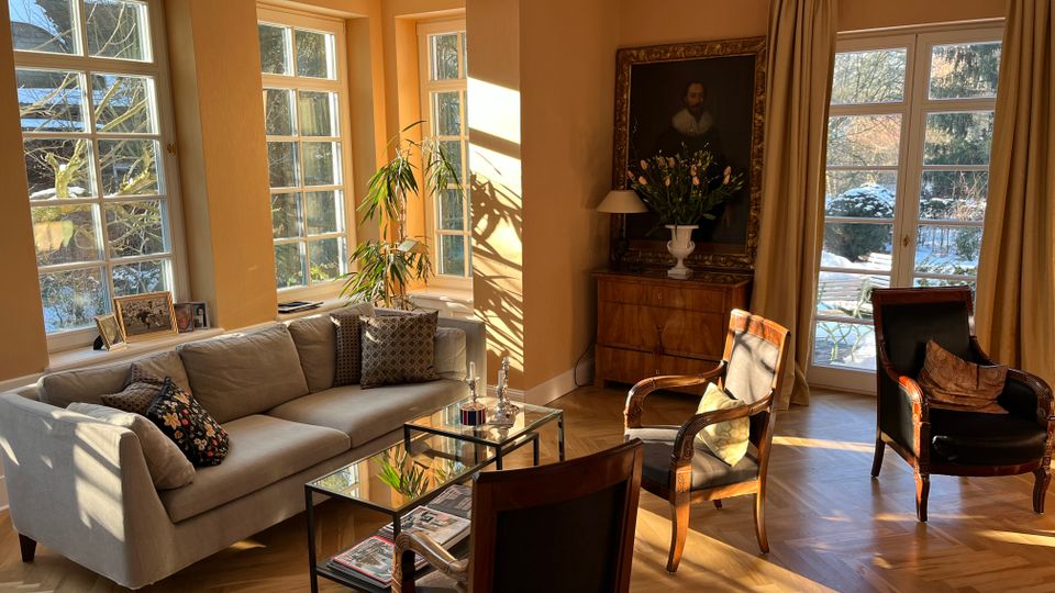 Helles Wohnzimmer mit hohen Sprossenfenstern und einer verglasten Terrassentür, die viel Sonnenlicht auf den Fischgrätparkettboden fallen lässt. Im Raum stehen ein graues Ecksofa, zwei Glas-Couchtische, mehrere klassische Sessel sowie eine Kommode mit großem gerahmtem Porträt und Vorhängen an der Fensterfront.