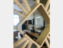 Blick durch ein Gitter in einen kleinen, altmodischen Büroraum mit gemusterter Tapete und Fliesenboden. Entlang der Wand stehen Schreibtische mit Telefon und technischen Geräten; daneben eine Glastür mit Fluchtwegzeichen und ein großes Fenster mit Vorhang.