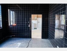 Innenraum mit glänzenden schwarzen Kacheln an Wänden und Boden, links ein schmaler hoher Spiegel und rechts eine spiegelnde Wandfläche. In der Mitte führt eine Türöffnung in ein helles, weiß gefliestes Badezimmer mit Dusche und kleinem Spiegel; auf dem Boden liegt ein Kabel.