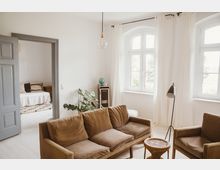 Helles Wohnzimmer mit braunem Sofa und Sessel, daneben ein kleiner runder Korbtisch. Im Hintergrund ist ein abgetrennter Raum mit Bett sichtbar, große Fenster mit weißen Vorhängen lassen viel Tageslicht hinein.