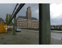 Backsteingebäude mit einem zentralen Uhrturm und zahlreichen weißen Fenstern, umgeben von einer gepflasterten Fläche mit parkendem Auto, Containern und Metallträgern im Vordergrund; im Hintergrund ein bewölkter Himmel.