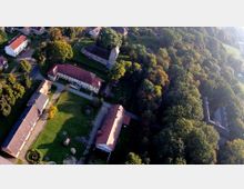 Luftaufnahme eines ländlichen Anwesens mit mehreren Gebäuden, darunter ein rechteckiges Hauptgebäude, Nebengebäude mit roten Ziegeldächern und eine kleine steinerne Kirche. Die Umgebung ist geprägt von weitläufigem Grün, Bäumen und angrenzendem Wald.