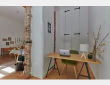 Ein minimalistisches Arbeitszimmer mit einer Holztischplatte auf Metallfüßen, einem grünen Stuhl und einem Laptop. Im Hintergrund sind freiliegende Ziegelwände, ein weißer Wandschrank mit schwarzen Beschlägen und ein Bett in einem angrenzenden Raum zu sehen, dekoriert mit Wandbildern.