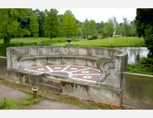Halbrunde steinerne Bank mit verzierten Seitenornamenten und einem dekorativen Bodenmosaik, gelegen in einer Parklandschaft mit Rasenflächen, Bäumen und einem Teich im Hintergrund.