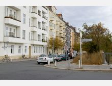 Straßenszene mit mehrstöckigen Wohngebäuden in hellen Farben, Balkonen und vereinzelten Pflanzen. Im Vordergrund parken Autos entlang der Straße, ein Verkehrsschild weist auf eine Tempo-30-Zone hin, und rechts stehen Bäume und trockene Büsche entlang einer Brücke mit dem Straßenschild „Monumentenbrücke“.