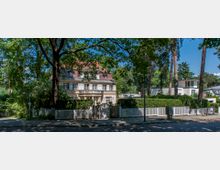 Zweistöckige Villa mit heller Fassade und roten Dachschindeln, umgeben von einem Garten mit hohen Bäumen und einer weißen Zaunanlage, gelegen in einer grünen Wohngegend mit Kopfsteinpflasterstraße im Vordergrund.