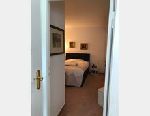 Blick durch eine offene Tür in ein Schlafzimmer mit einem Doppelbett, das eine helle, gemusterte Bettdecke hat. An den Wänden hängen zwei kleine, gerahmte Bilder, und neben dem Bett steht ein weißer Nachttisch mit einer Lampe.