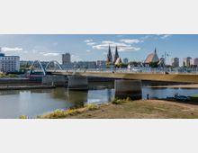 Flusslandschaft mit einer modernen Straßenbrücke im Vordergrund, die auf Betonpfeilern ruht. Im Hintergrund sind Kirchtürme, Wohngebäude und ein Parkbereich zu sehen, bei klarem Himmel und hellem Tageslicht.