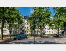 Straßenkreuzung in einem Wohngebiet mit Straßenschildern "Liebenowzeile" und "Denkstraße", umgeben von mehrstöckigen, beige gehaltenen Wohngebäuden und Bäumen, die Schatten spenden. Im Vordergrund sind parkende Autos und eine asphaltierte Straße zu sehen.