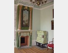 Historischer Raum mit einem dekorativen Kamin in grünlicher Wandfarbe, flankiert von rosafarbenen Säulen und einem Gemälde mit Landschaftsszene. Rechts daneben steht ein grün-gelb gemusterter Kachelofen, und im Vordergrund sind Holzfußboden sowie ein Kronleuchter sichtbar.