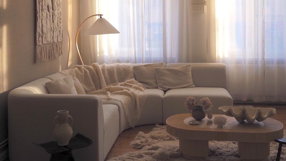 Sunlit modern living room with a white sectional sofa, textured throw blankets and pillows, and an arched floor lamp beside large windows with sheer curtains. A low oval wooden coffee table sits on a shag rug, with neutral-toned decor and a woven wall hanging on the left wall.