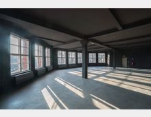 Leerer, modern-industrieller Raum mit großen Fenstern, schwarze Wände und sichtbaren Stahlträgern. Tageslicht fällt durch die Fenster und erzeugt lange Schatten auf dem Betonboden.