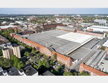 Luftaufnahme eines weitläufigen Gewerbe- und Industriekomplexes mit roten Ziegelgebäuden im historischen Stil und großen Flachdachhallen. Im Vordergrund befinden sich Straßen mit Bäumen und vereinzelten Wohngebäuden; im Hintergrund erstreckt sich eine städtische Landschaft mit Wohnhäusern und Grünflächen.