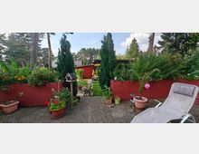 Gartenbereich mit bunten Blumen und Pflanzen in roten Holzpflanzkästen, dahinter eine Rasenfläche mit Gartenbank und einem roten Gartenhaus. Im Vordergrund ein gepflasterter Sitzbereich mit Blumentöpfen, einer Liege und einem Vogelfutterhaus.