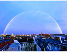 Ansicht eines städtischen Dachs mit Ziegel- und Flachdächern, über denen ein vollständiger Regenbogen vor einem bläulichen Himmel sichtbar ist. Im Hintergrund sind Kirchtürme und andere markante Gebäude einer Stadtsilhouette zu erkennen.