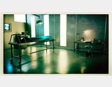 Ein steriler Raum mit einem Obduktionstisch in der Mitte, abgedeckt mit einem blauen Tuch, beleuchtet von einer Deckenlampe. An der Seite steht ein Schreibtisch mit einer Lampe, einem Bürostuhl und verschiedenen Geräten; im Hintergrund leuchten vertikale Lichtelemente.