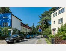 Wohnsiedlung mit mehrfarbigen Reihenhäusern entlang einer ruhigen, von Autos gesäumten Straße; links ein blaues Haus, rechts gelbe Fassaden, im Hintergrund Bäume und ein Straßenschild für eine Sackgasse.