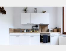 Moderne Küche mit weißen Schränken und einer Arbeitsplatte in Holzoptik. Sichtbar sind ein Waschbecken, ein Herd mit Backofen, verschiedene Kochutensilien, Obst auf der Arbeitsfläche sowie ein großes Fenster rechts.