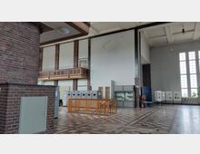 Innenraum eines historischen Gebäudes mit hohen Decken, großen Fenstern und gemustertem Fliesenboden. Zu sehen sind eine Holzbalustrade auf einer Galerie, Schautafeln, Vitrinen sowie technische Schaltkästen entlang der Wände.