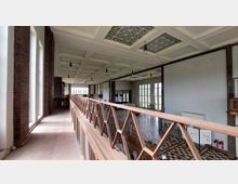 Innenansicht eines großen Saals mit hoher Decke und Kassettierung, großen Fenstern auf der rechten Seite und roten Backsteinwänden links. Ein hölzernes Geländer im Vordergrund grenzt eine Art Galerie oder Laufsteg ab, der über den Raum führt. Der Boden ist mit einem geometrisch gemusterten Fliesenboden versehen.