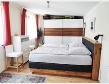 Schlafzimmer mit einem Doppelbett mit Holzrahmen und dunkler Polsterung, gegenüber einem großen Kleiderschrank mit weißen und holzfarbenen Paneelen. Links stehen eine weiße Kommode, zwei Stühle und eine Topfpflanze, mit roten Vorhängen an den Fenstern.