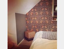 Schlafzimmer mit schräge Decke, einer Wand mit floralem Tapetenmuster in warmen Tönen, einem geflochtenen Würfelhocker und einem Bett mit weißer Tagesdecke sowie gemustertem Überwurf. Ein Regal mit Pflanzen und eine Lampe sind im Hintergrund sichtbar.