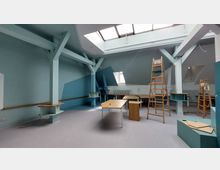 Ein heller Raum mit blauen Wänden und einer Dachschräge, der durch ein großes Oberlicht beleuchtet wird. Im Raum stehen Holztische, Leitern und Regale; freiliegende Holzbalken und Heizkörper an den Wänden sind sichtbar.