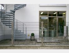 Eine Außentreppe aus Metall in einer Wendelform vor einer weißen Fassade, daneben ein großes Fenster mit Blick auf industrielle Geräte im Inneren. Im Vordergrund steht ein Metallzaun, hinter dem ein kleiner Kübel mit einer Pflanze auf Rollen platziert ist.