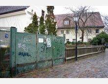 Eine gepflasterte Straße führt zu einem alten Haus mit heller Fassade und roten Dachziegeln, umgeben von Bäumen. Im Vordergrund befindet sich ein grünes, mit Graffiti bemaltes Metalltor und ein Holzzaun neben einer Laterne.
