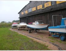 Holzgebäude mit dunkler Fassade, an dem gelbe Schilder mit Hinweisen wie „Ankauf Verkauf“ und „Service Center“ angebracht sind. Davor stehen mehrere kleine Motorboote auf Trailern, während im Hintergrund Wiese und Sträucher sichtbar sind.