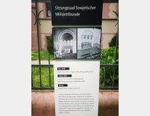 Informationsschild vor einem historischen Gebäude mit der Aufschrift „Sitzungssaal Sowjetischer Militärtribunale“. Darunter befinden sich zwei historische Schwarz-Weiß-Fotos des Innenraums sowie Text, der die Nutzung der Kapelle vor 1945, während der Sowjetzeit und heute erklärt. Im Hintergrund sind Teile des rosafarbenen Gebäudes und eine Grünfläche sichtbar.