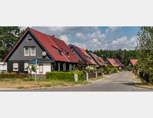 Eine ruhige Wohnstraße mit Einfamilienhäusern im Stil von Satteldachhäusern, die rote und graue Dachziegel haben. Die Straße ist von gepflegten Vorgärten und Hecken gesäumt und wird von Bäumen in der Ferne umgeben.