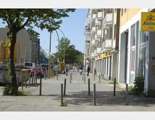 Straßenszene in einer Stadt mit breitem Gehweg, rechts Wohn- und Geschäftshäuser mit Balkonen und Schaufenstern, links parkende Autos sowie eine Baumreihe. Im Hintergrund ist ein Schild einer U-Bahn-Station und weiteres Stadtverkehr zu sehen.