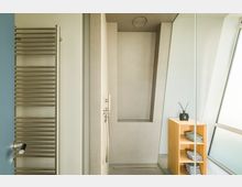 Modernes Badezimmer mit einer offenen, wandintegrierten Dusche und einer Überkopfdusche. Links ein Heizkörper in Handtuchhalter-Optik, rechts ein Holzregal mit Handtüchern und einer Duftflasche; ein großes Fenster sorgt für viel Tageslicht.
