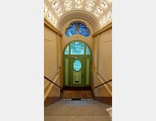 Treppenhaus mit einer grünen Holztür und ovalem Fenster im Eingangsbereich. Über der Tür befindet sich ein buntes Glasfenster mit abstraktem Motiv, während die Raumdecke mit einem weißen Gewölbe und vielen kleinen Leuchten verziert ist.