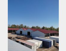 Großer Lagerhof mit mehreren weißen Hallenbauten mit roten Dächern, umgeben von Bäumen im Hintergrund. Im Vordergrund sind gestapelte Frachtcontainer sichtbar.