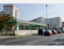 Eine städtische Straßenansicht mit dem Eingang zur U-Bahn-Haltestelle „U-Bhf Innsbrucker Platz“, gekennzeichnet durch ein grünes, gläsernes Überdach. Im Hintergrund sind mehrstöckige Wohn- und Bürogebäude zu sehen, während Autos auf der Straße im Vordergrund fahren.