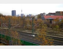 Blick auf Bahngleise mit Oberleitungsanlagen im Vordergrund, umgeben von herbstlichen Bäumen. Im Hintergrund sind Stadthäuser, Industriegebäude und der Berliner Fernsehturm sichtbar.