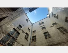 Blick von unten in einen engen Innenhof mit hohen Gebäudewänden aus weißen Ziegeln und mehreren rechteckigen Fenstern. In der Mitte ist ein Stück blauer Himmel sichtbar.