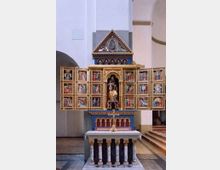 Altartafel in einer Kirche mit geöffnetem Flügelaltar, der zahlreiche religiöse Gemälde zeigt. In der Mitte befindet sich eine zentrale Figur unter einer kunstvoll gestalteten Spitze, darunter ein Sockel mit mehreren Säulen.