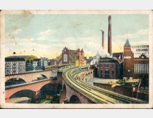 Historische Stadtansicht mit Eisenbahnbrücke und Industriebauten, darunter Backsteingebäude mit Schornsteinen, Beschriftungen wie „Kühlhaus“ erkennbar; im Hintergrund Wohnhäuser und eine Markthalle.