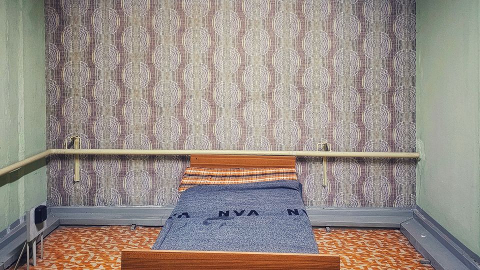 Kleiner, spartanischer Schlafraum mit Einzelbett aus Holz vor einer gemusterten Tapetenwand. Auffällig sind der orange-braun gemusterte Linoleumboden, grün gestrichene Seitenwände sowie große Lüftungs- und Rohrleitungen unter der Decke.