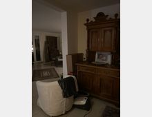 Wohnzimmer im gedämpften Licht mit einem großen, dunkelbraunen Holzschrank an der Wand, auf dem Bilderrahmen und Dekorationen stehen. Im Vordergrund ein beigefarbener Sessel mit einer dunklen Decke, daneben ein Staubsauger; im Hintergrund ein Esstisch, Teppiche und Glastüren.