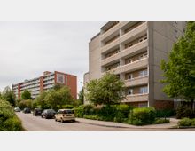 Wohngebäude in einer städtischen Umgebung mit mehreren Etagen und Balkonen, flankiert von Bäumen und Sträuchern. Im Vordergrund verläuft eine Straße mit geparkten Autos, umgeben von Grünflächen.