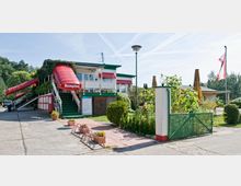 Ein kleiner, traditioneller Empfangsbereich mit rotem Vordach und der Aufschrift „Rezeption“, ergänzt durch grüne Treppen und Pflanzen ringsum. Im Hintergrund sind weitere flache Gebäude und ein gepflegter Garten mit Zaun sowie eine Flagge sichtbar.