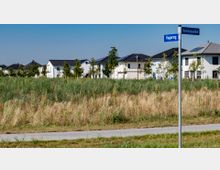 Eine Wohnsiedlung mit modernen weißen Einfamilienhäusern und grauen Dächern, umgeben von einer Wiese mit hohem Gras. Im Vordergrund ein Weg und ein Straßenschild mit den Namen "Hugoring" und "Rousseauallee".
