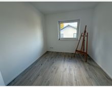 Kleiner, heller Raum mit weiß gestrichenen Wänden und einem Fenster, durch das ein Teil eines Hausdaches sichtbar ist. Der Boden besteht aus hellem Holzimitat, und in einer Ecke steht eine leere Holzstaffelei.