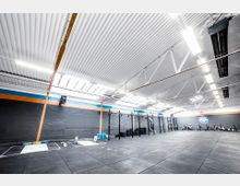 Große, modern gestaltete Sporthalle mit einem hohen Deckenbereich aus Metallpaneelen und sichtbarer Stahlträgerkonstruktion. Der Raum enthält Trainingsbereiche mit schwarzen Fitnessgeräten, Gewichten, Bodenmatten und einer dunklen Wand mit orangefarbenen Akzenten.