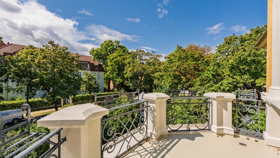 Blick von einem hellen Balkon mit dekorativem schmiedeeisernem Geländer und massiven Eckpfeilern auf eine von hohen Bäumen gesäumte Wohnstraße. Im Hintergrund sind mehrstöckige Wohnhäuser mit roten Ziegeldächern unter blauem Himmel zu sehen.